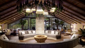 A spacious living area with a large, circular sofa, a central wooden fire pit, and hanging plants. - Hotel Quelle Nature Spa Resort (Valle di Casies)