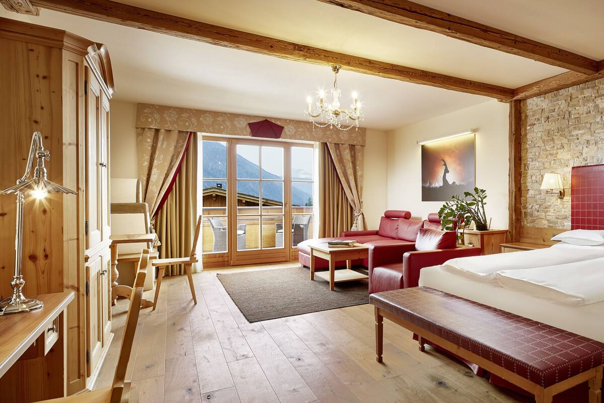 Ein Hotelzimmer mit einem großen Bett, einer Bank, einem Schreibtisch, einem Sessel, einem Tisch und Blick auf Berge.