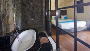 A bathroom with a stone wall, a bathtub, and a shower. There is a bed with white bedding and blue pillows in the adjacent room. - Alam Bali Hotel (Nusa Dua)