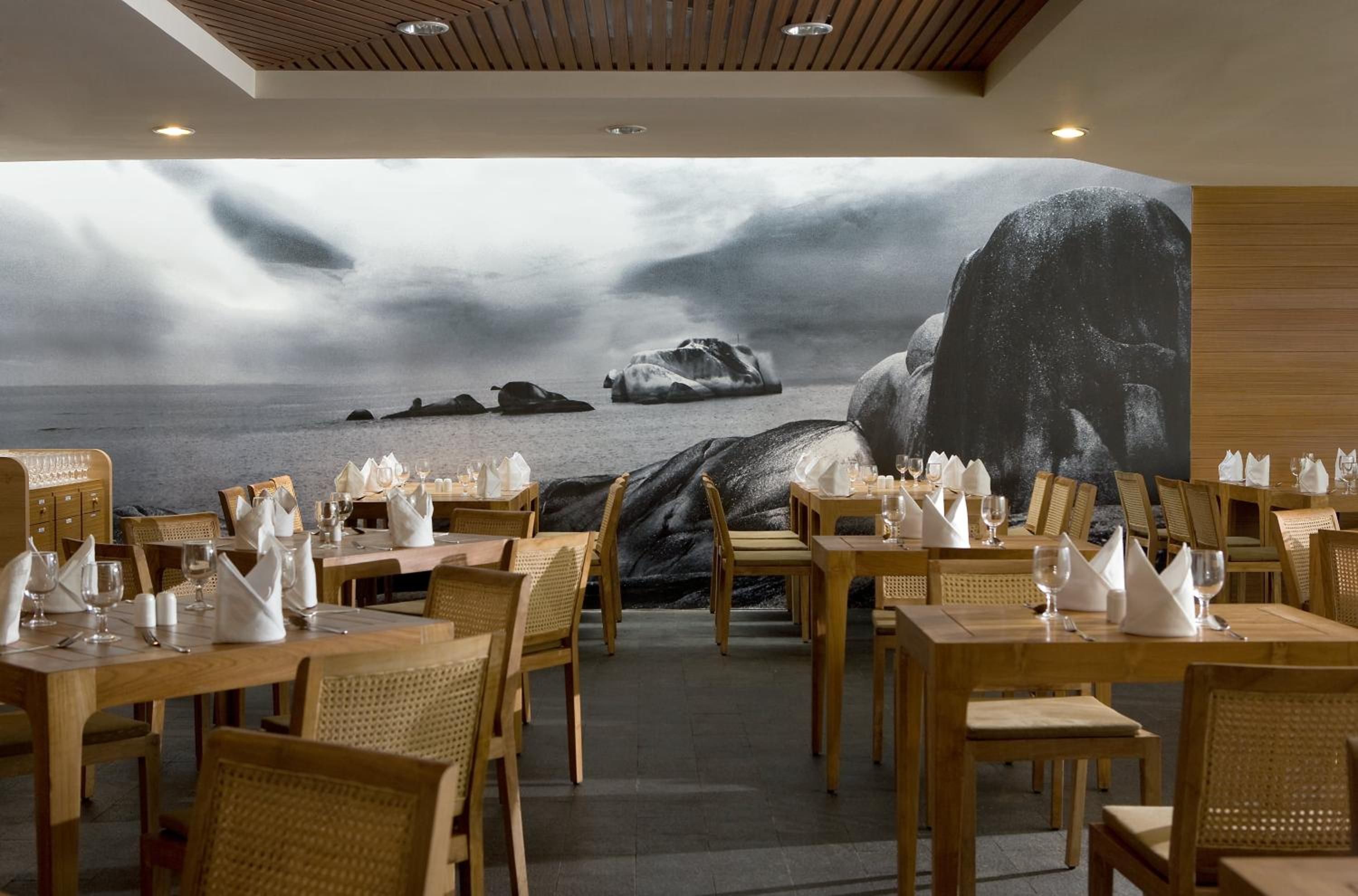 Een restaurant met houten tafels en stoelen, een groot muurschildering van een rotsachtig landschap met een waterpartij, en een plafond met houten latten en inbouwverlichting.