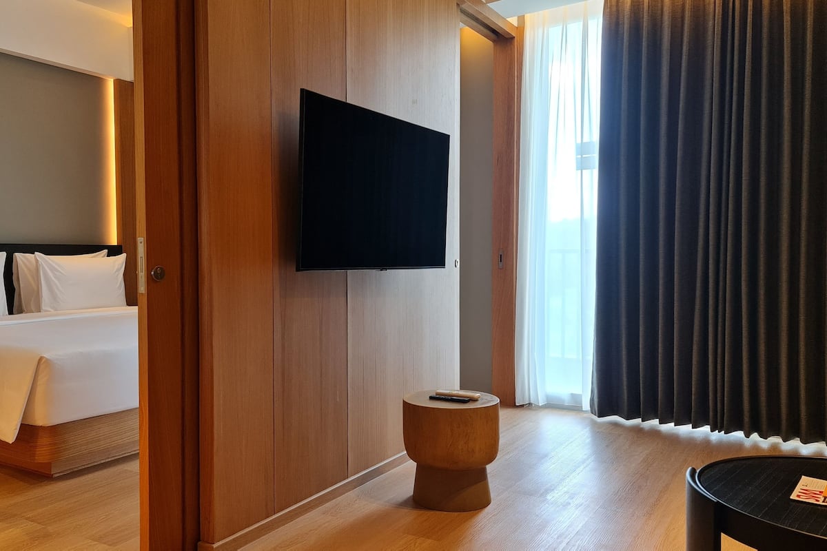 Una habitación de hotel con cama, televisión montada en la pared, piso de madera y ventana con cortinas.