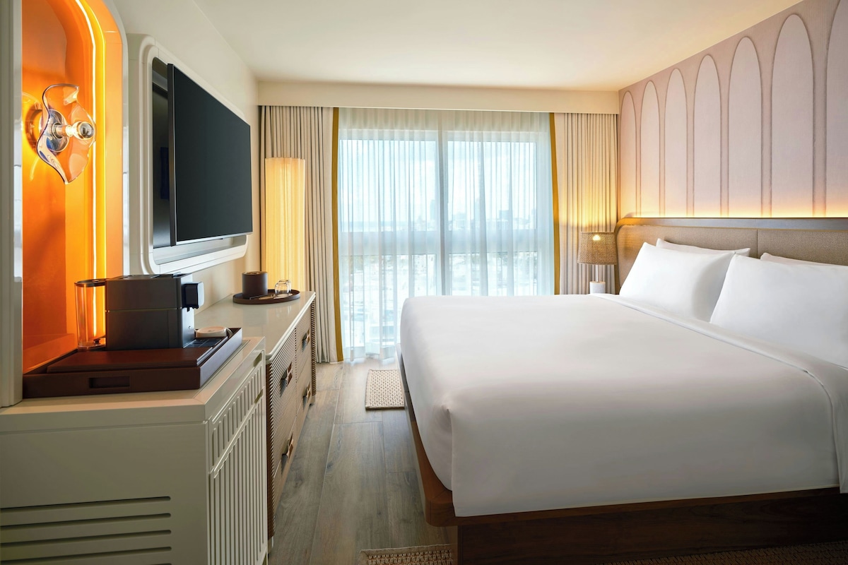 Habitación de hotel con una cama grande, un televisor de pantalla plana, una cafetera y vistas de la ciudad a través de la ventana.