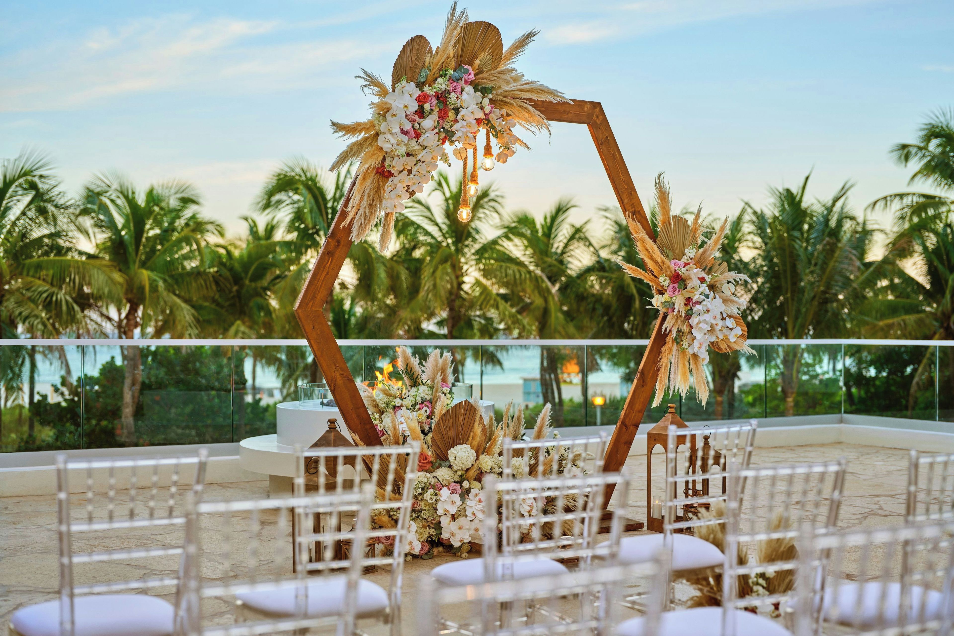 An outdoor wedding setup with a wooden arch adorned with flowers and pampas grass, white chairs arranged in rows, and a view of palm trees and a beach in the background.
