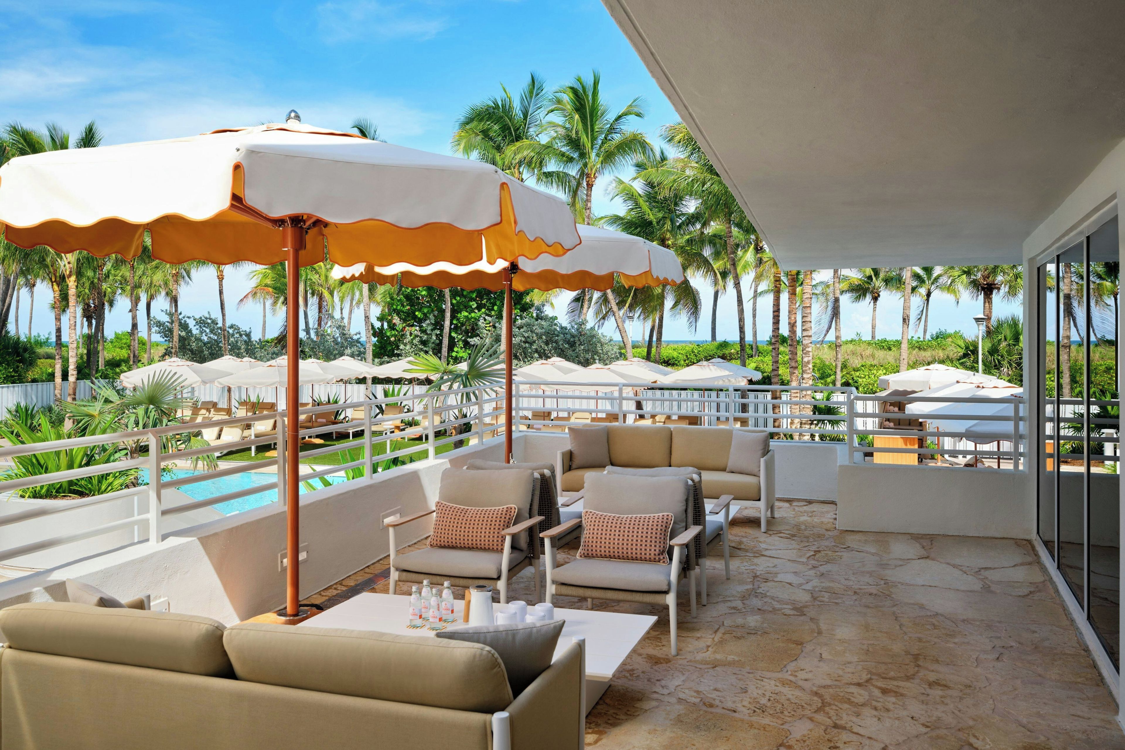 A sunlit outdoor seating area with lounge chairs and umbrellas, overlooking a pool and palm trees.