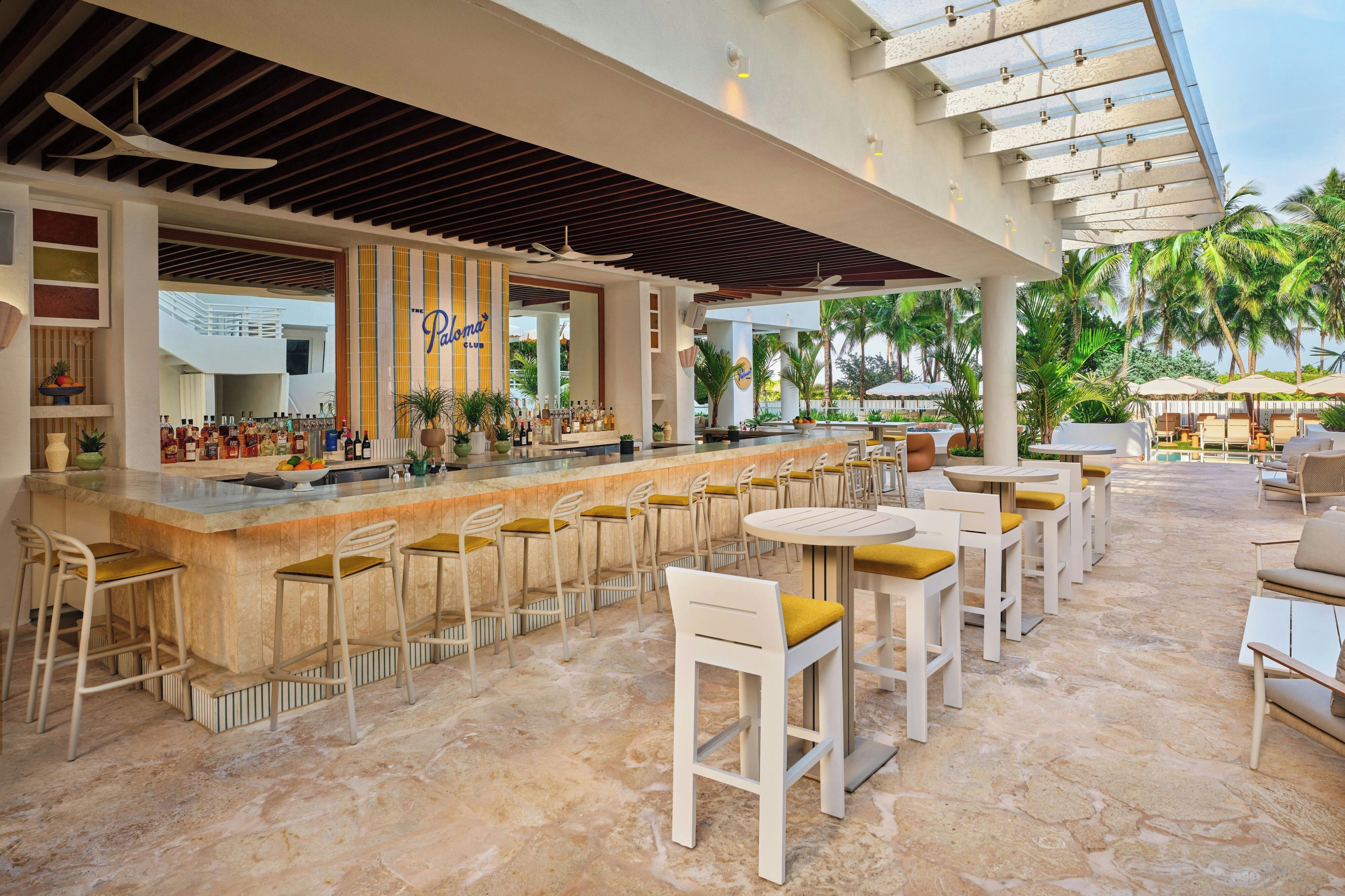 Un espace bar avec un comptoir, des tabourets et des tables, entouré de palmiers et d’une piscine.