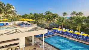 Un área de piscina con sillones reclinables azules y sombrillas amarillas, rodeada de palmeras.