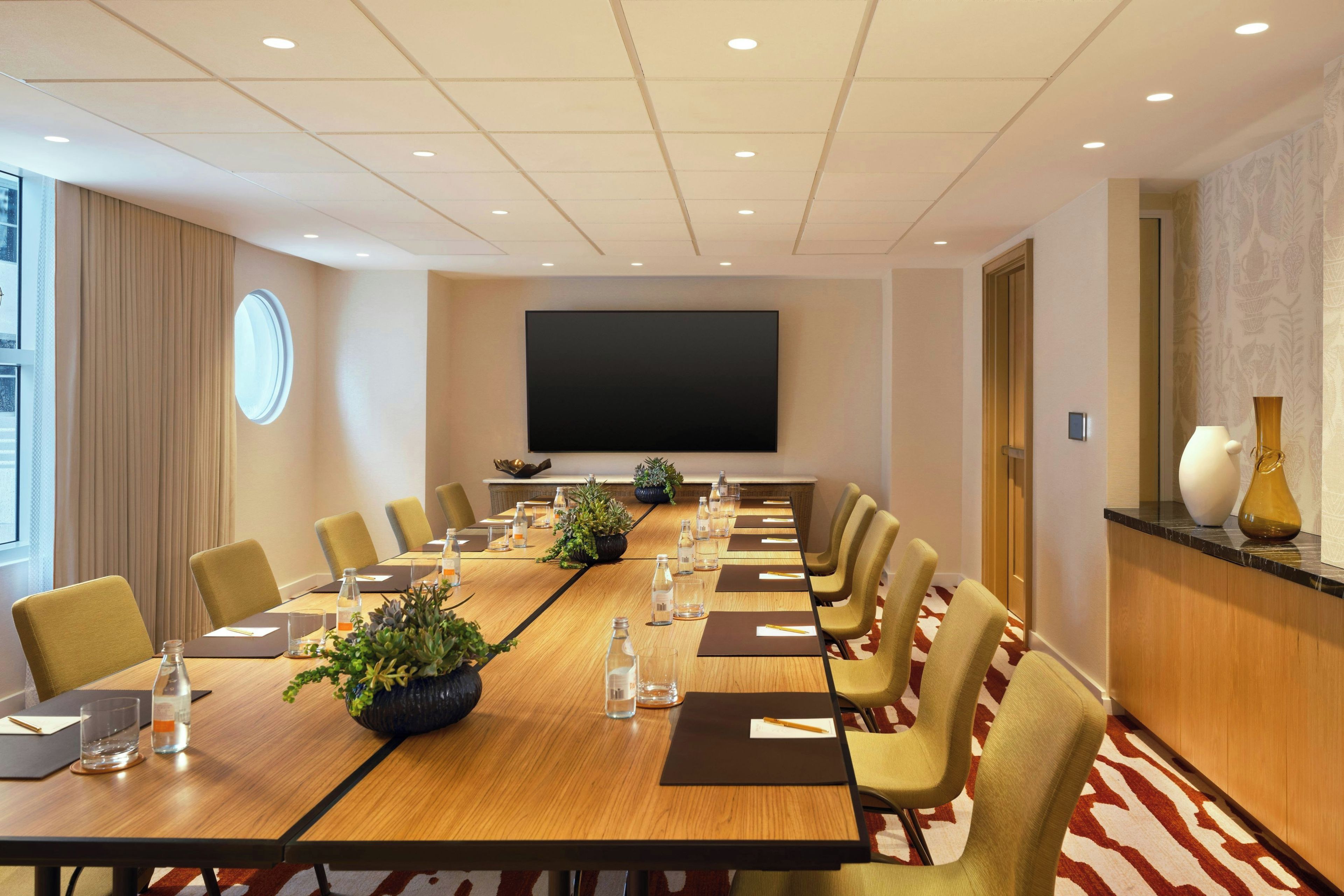 A conference room with a long table, chairs, a large screen, and a potted plant.