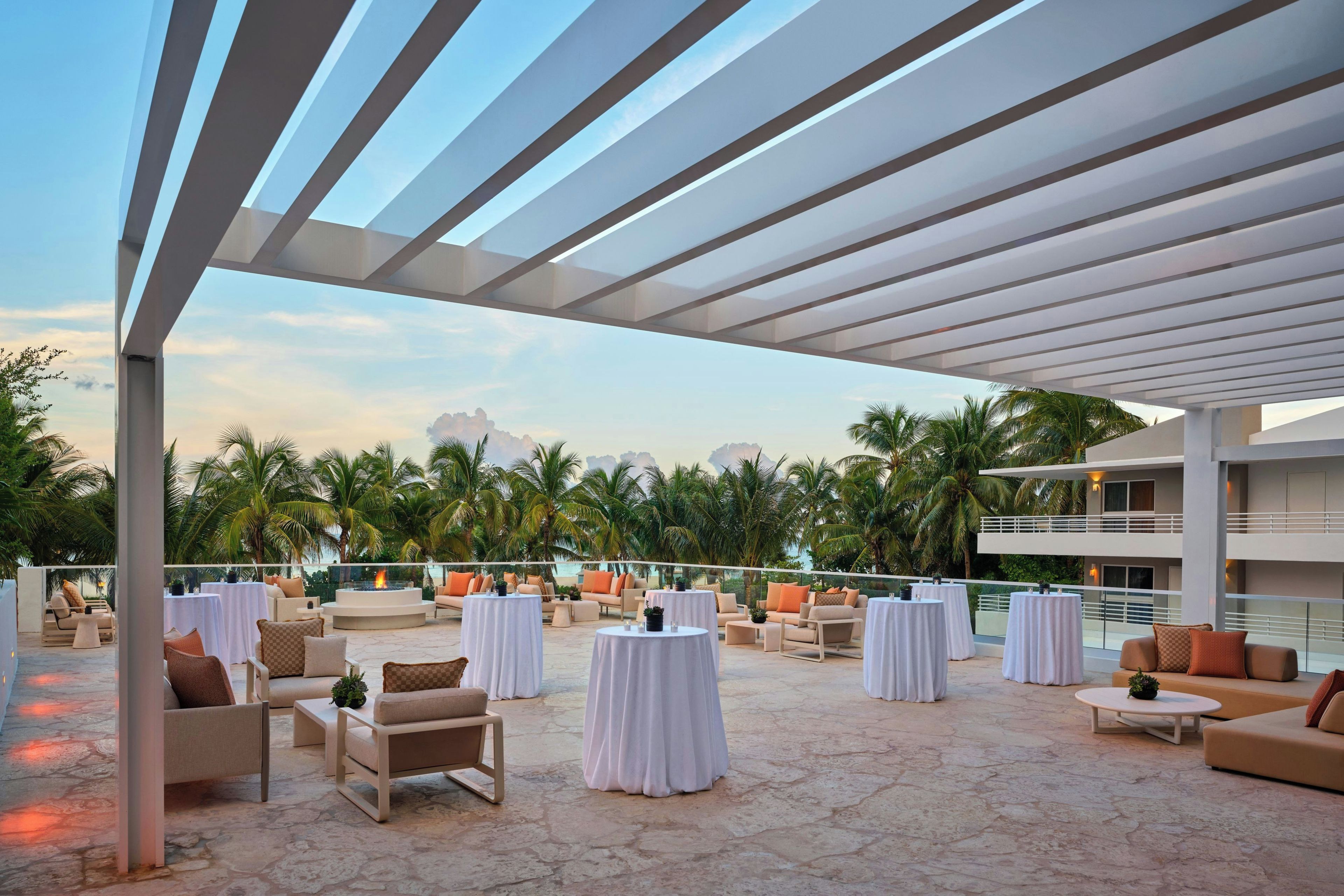 An outdoor patio with round tables and chairs, palm trees, and a view of the ocean.