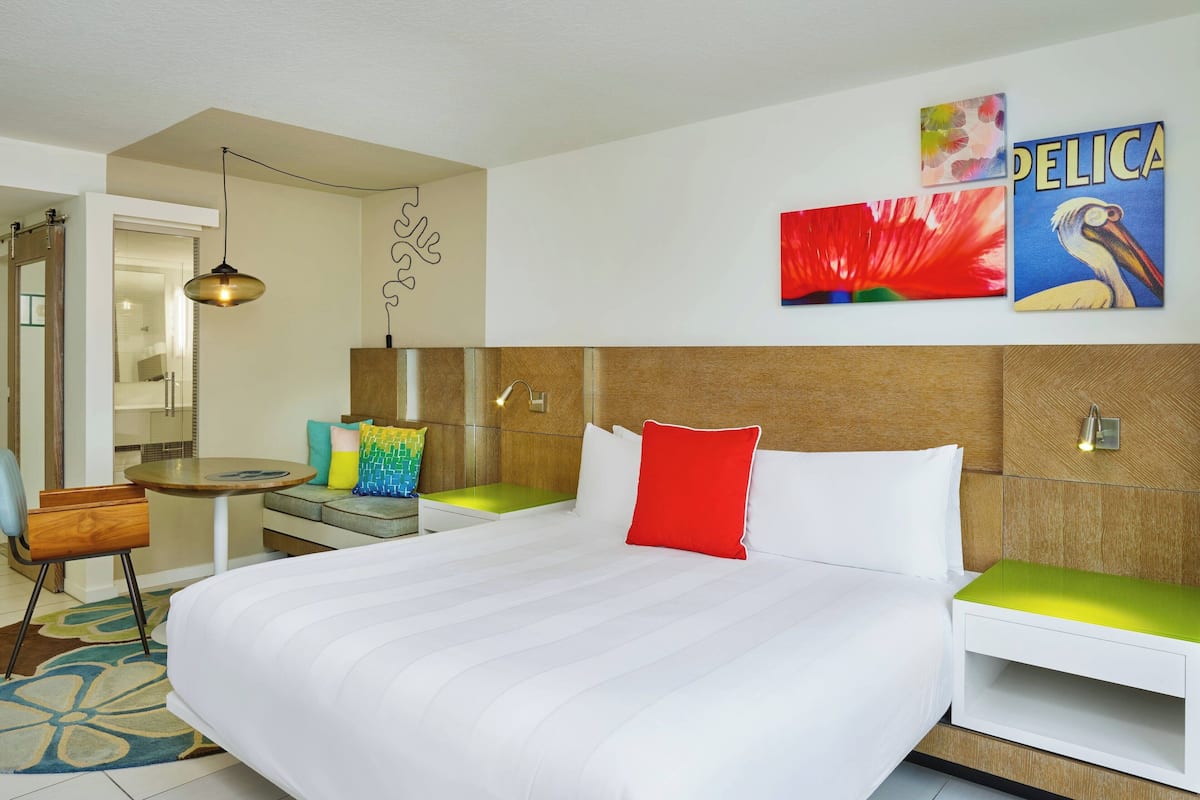 A hotel room with a large bed, a bedside table, a chair, a small table, and artwork on the wall.
