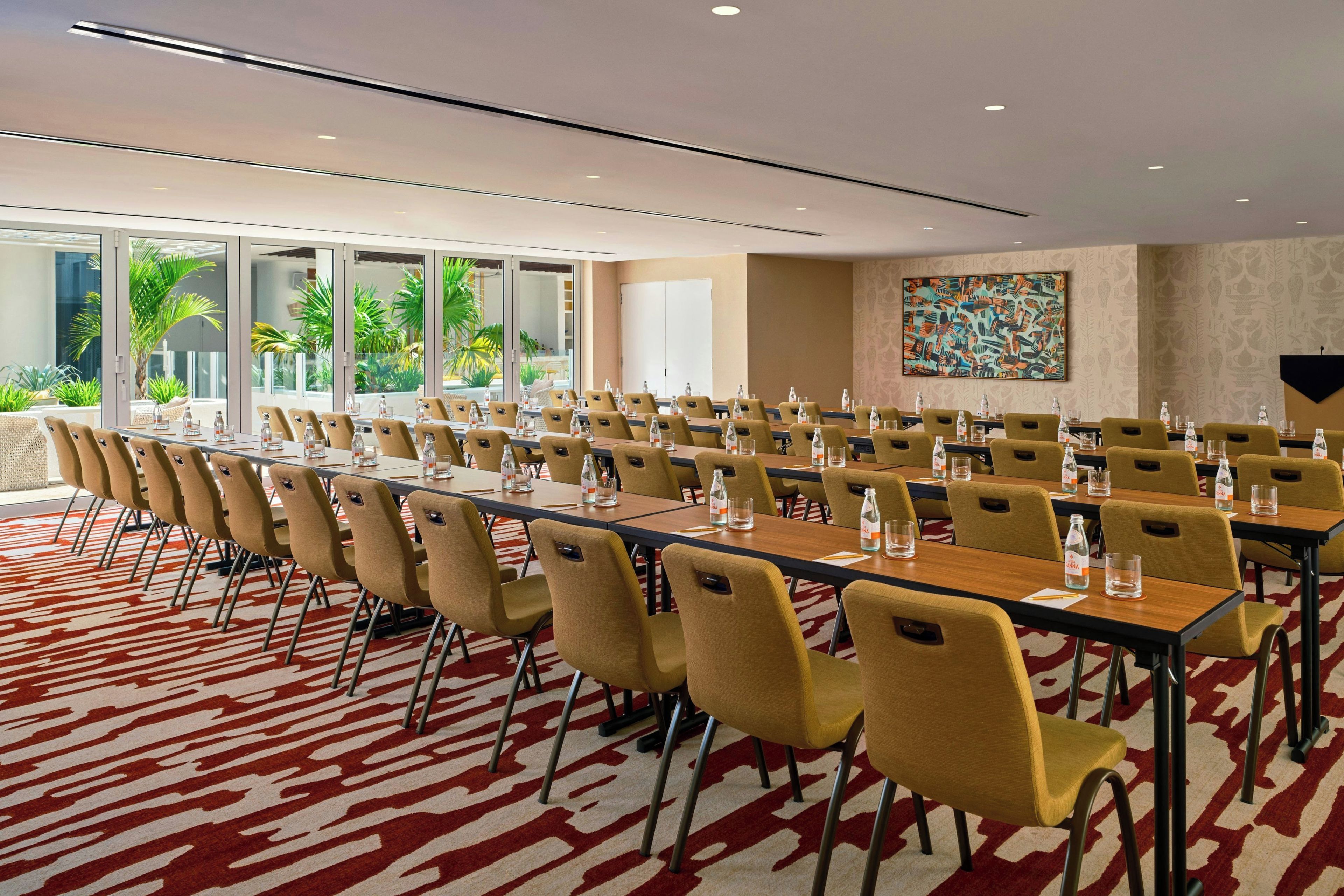 Ein Konferenzraum mit mehreren Tischen und Stühlen, großen Fenstern mit Blick auf Grünflächen und einem gemusterten Teppich.