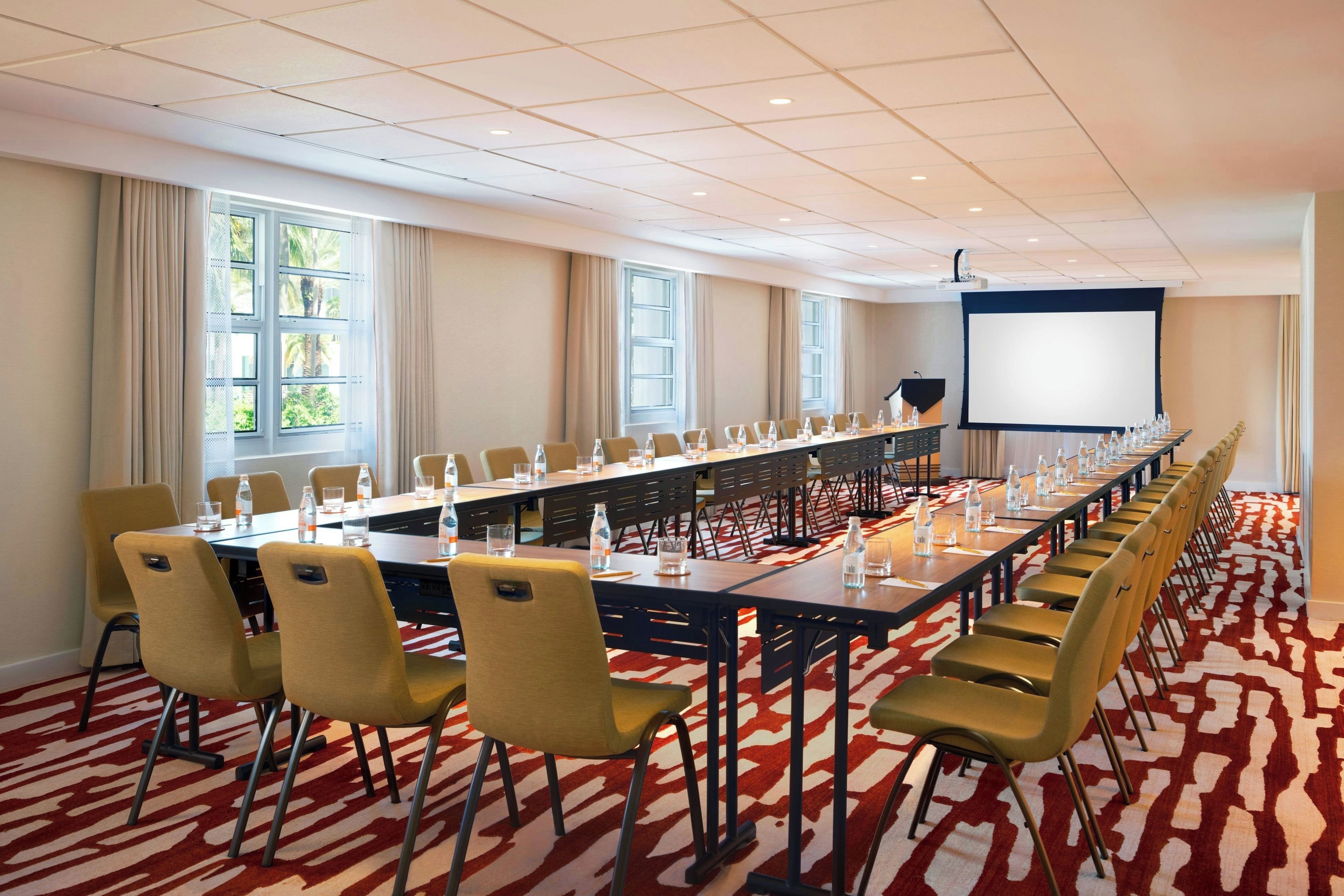 A conference room with a long table, chairs, a projector screen, and windows with curtains.