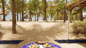 A beachside dining table with various dishes, surrounded by palm trees and a thatched-roof structure. - Blue Africa (Mbour)