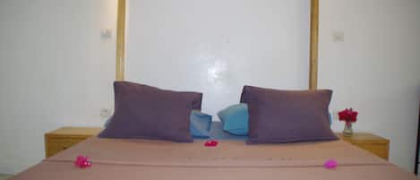 Una cama bien hecha con una colcha rosa y dos almohadas moradas, una mesita de noche con un florero y una pared blanca con una lámpara.