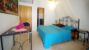 A room with a bed, a table with a chair, a painting on the wall, and a wooden door. - Blue Africa (Mbour)
