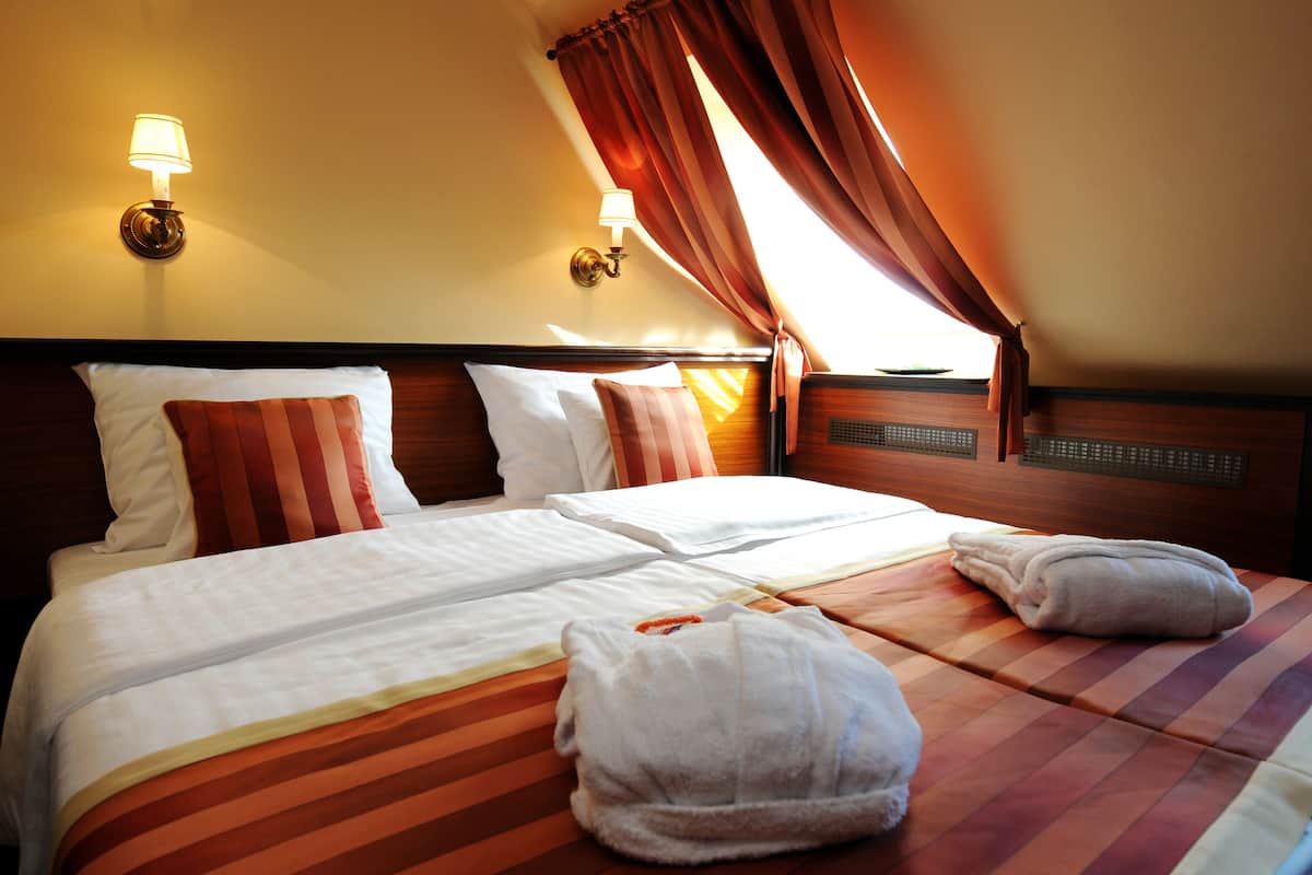 A hotel room with two beds, each with white linens and striped bedding. There are two wall-mounted lamps, a window with curtains, and a headboard with a built-in shelf.