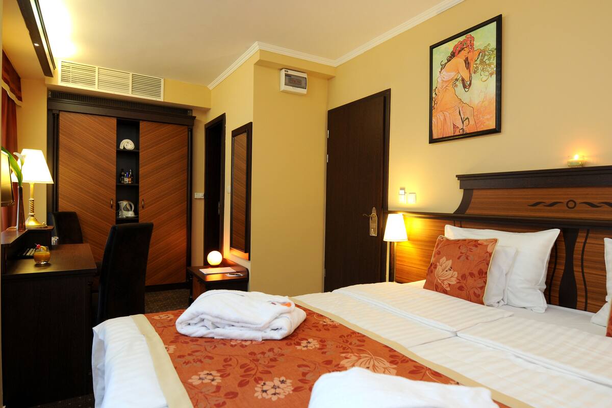A hotel room with a bed, a desk, a chair, a nightstand, a lamp, a painting on the wall, and a door.