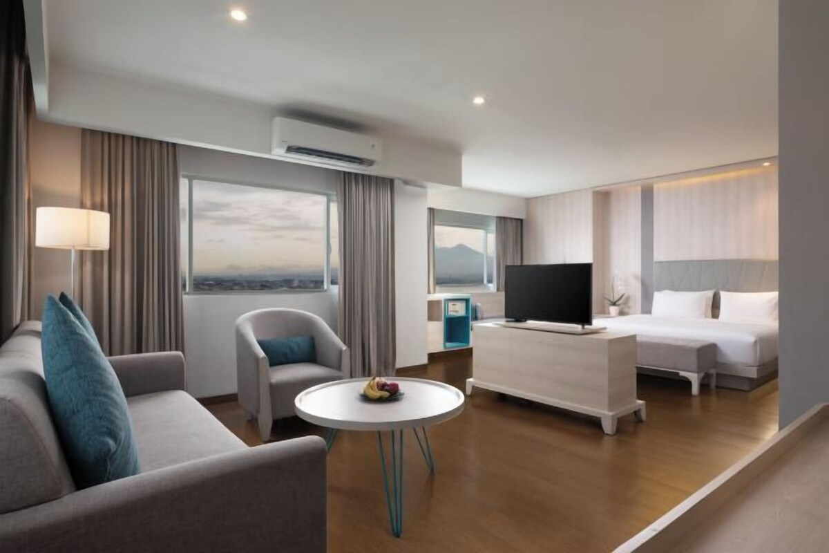 A modern hotel room with a sofa, a round table with fruit, a TV, a bed, and a city view.