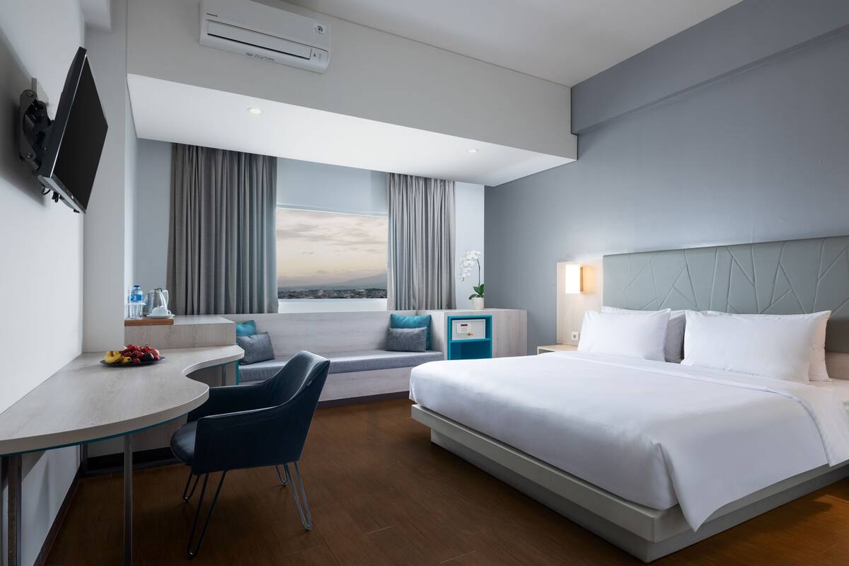 A modern hotel room with a bed, a desk, a chair, a window with curtains, and a TV mounted on the wall.