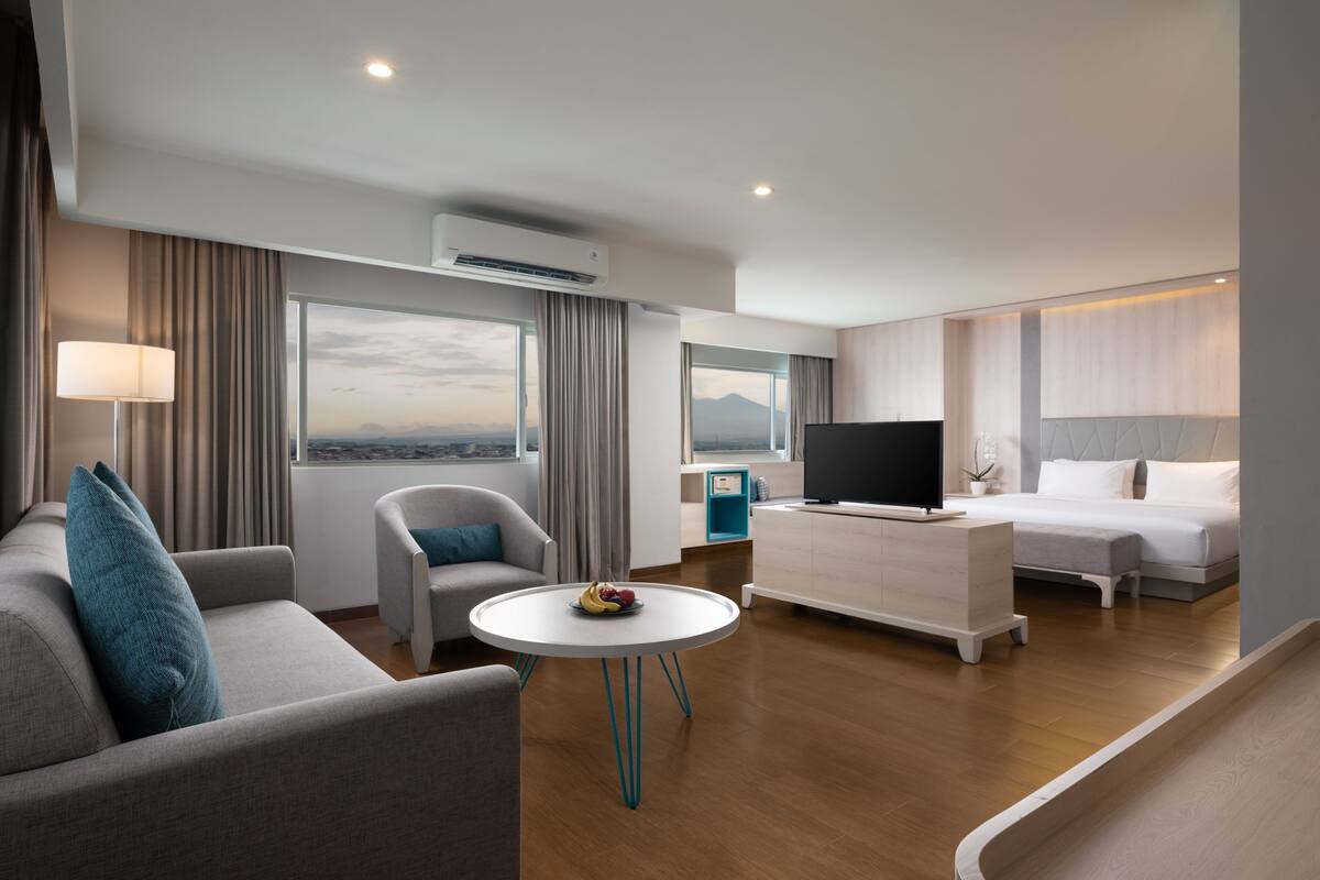 A modern hotel room with a sofa, armchair, round table, TV, and a bed.