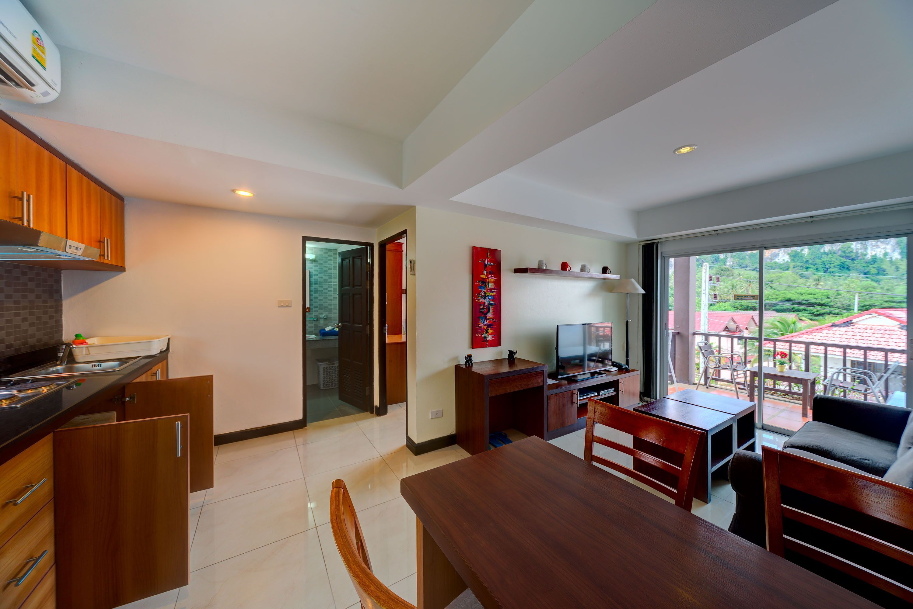 A modern kitchen with wooden cabinets, a dining area with a table and chairs, a TV, and a balcony with outdoor furniture.
