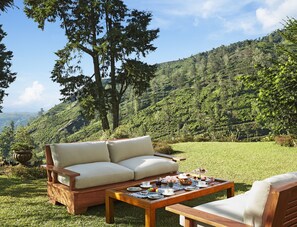 A wooden outdoor seating area with a sofa and a table set for a meal, surrounded by lush greenery and a hilly landscape.