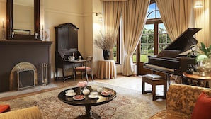 A formal living room with a grand piano, a fireplace, a dining table set for tea, and a large window with curtains.