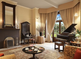 Un salón formal con un piano de cola, una chimenea, una mesa de comedor dispuesta para el té y un ventanal con cortinas.