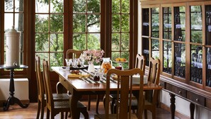A dining room with a wooden table set for six, surrounded by six chairs. Large windows provide a view of greenery outside.