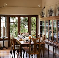 Comedor con una mesa de madera dispuesta para seis personas, rodeada por seis sillas. Amplias ventanas ofrecen vistas al verde exterior.
