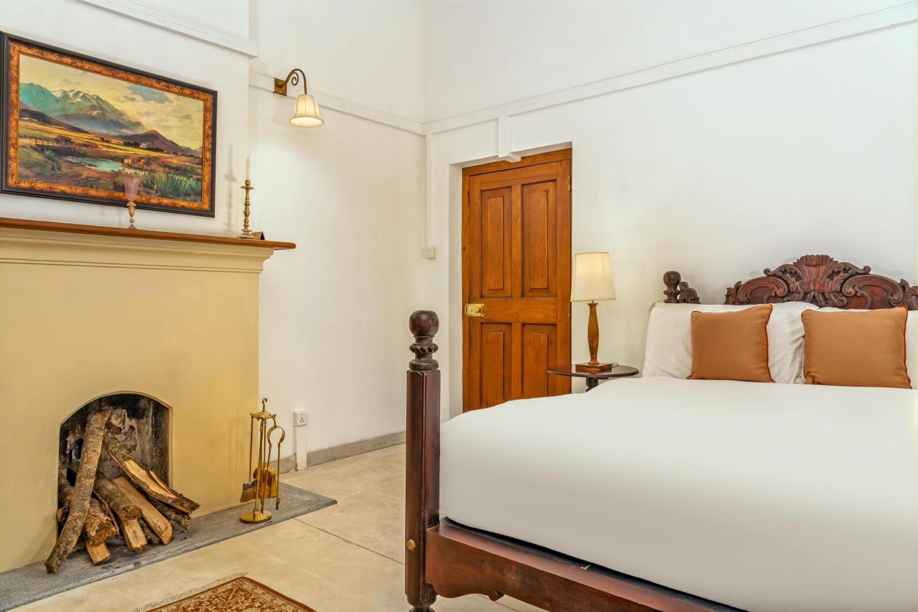 A bedroom with a large bed, a wooden door, a fireplace with logs, and a painting on the wall.