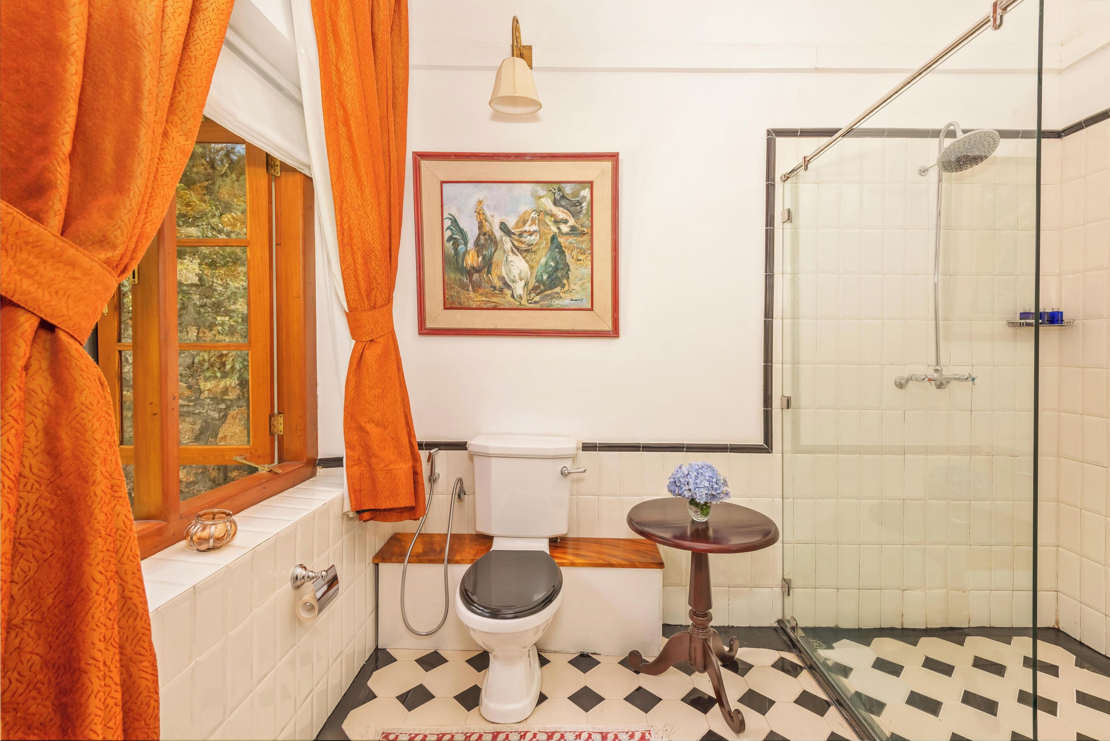 Un baño con inodoro, ducha, ventana con cortinas, un cuadro en la pared y una mesita con un florero.