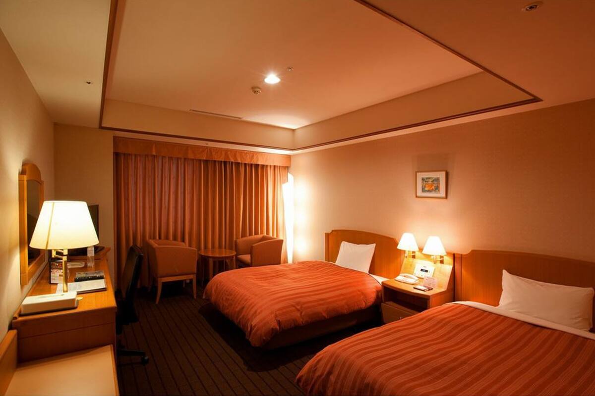 Hotellihuone, jossa on kaksi sänkyä, kirjoituspöytä lampun kanssa, tuoli ja pieni pöytä.