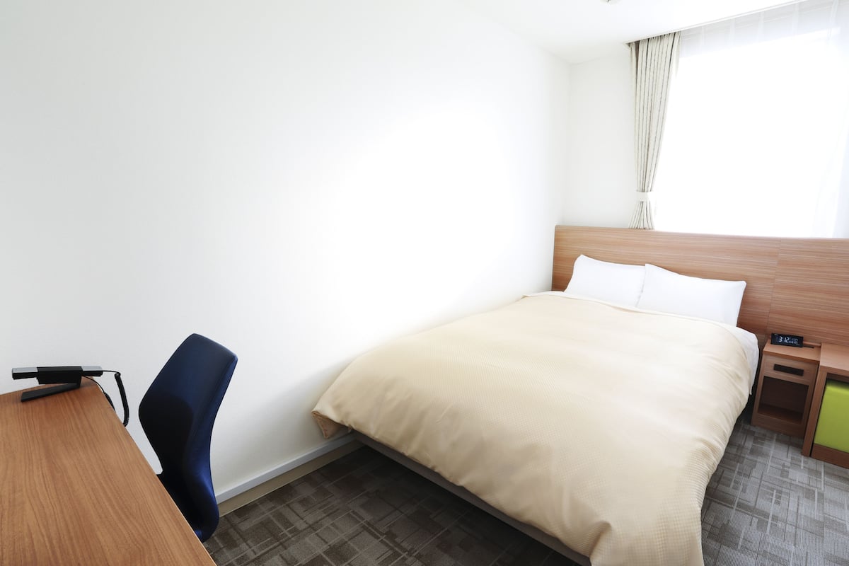 Una habitación de hotel con cama, escritorio, silla, mesita de noche y ventana con cortinas.