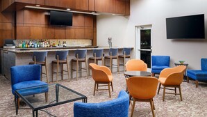 A bar area with wooden cabinets, a flat-screen TV, and bar stools. There are blue and orange chairs around a glass table.