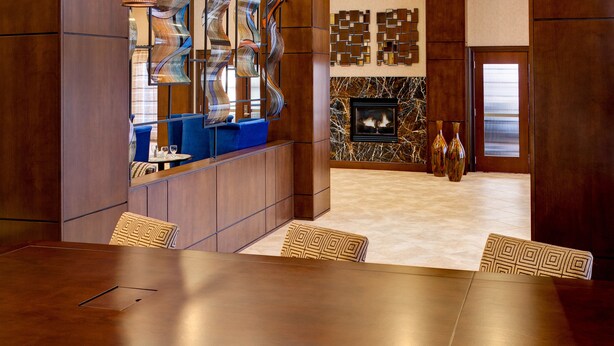 A modern hotel lobby with a large wooden reception desk, a fireplace, and a dining area with a long table and chairs.