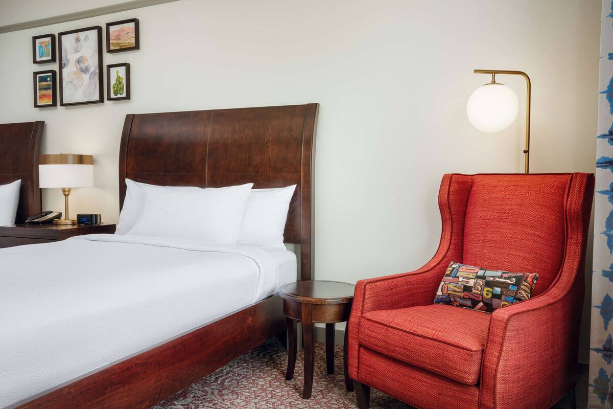 Hotellihuone, jossa on puinen sänky, punainen nojatuoli, pieni pöytä lampun kanssa ja seinä, jossa on kehystettyjä taideteoksia.