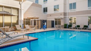 A swimming pool with lounge chairs and umbrellas, surrounded by a building with multiple windows and a tiled floor.