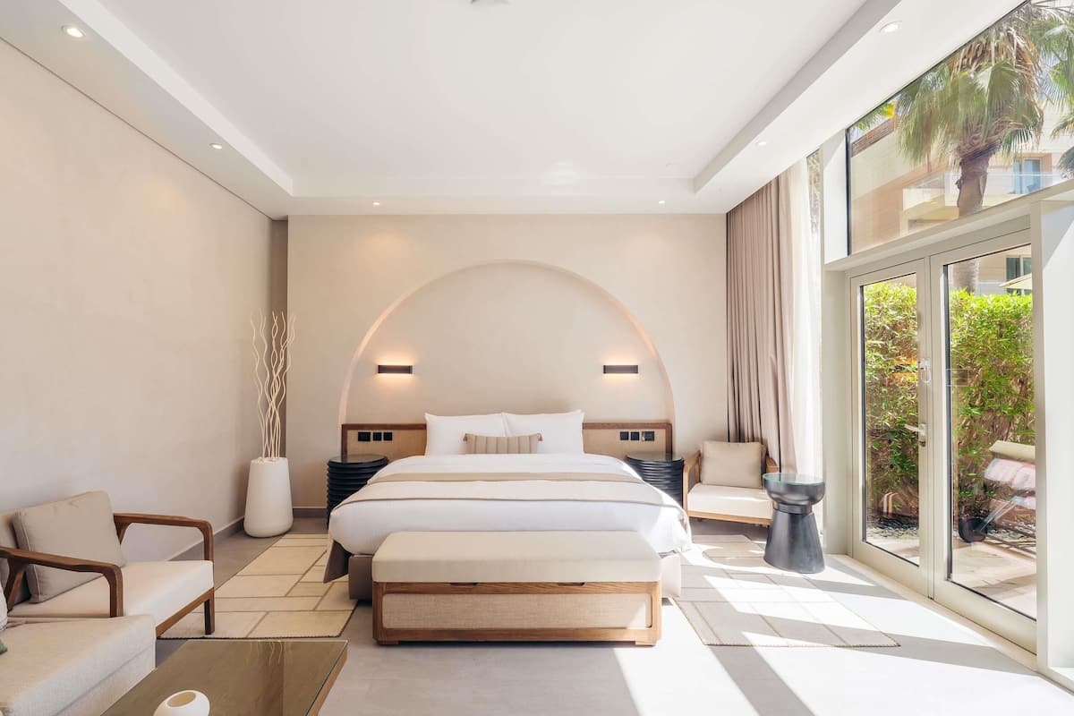 A modern hotel room with a large bed, a seating area with a coffee table, and a view of a garden through large windows.