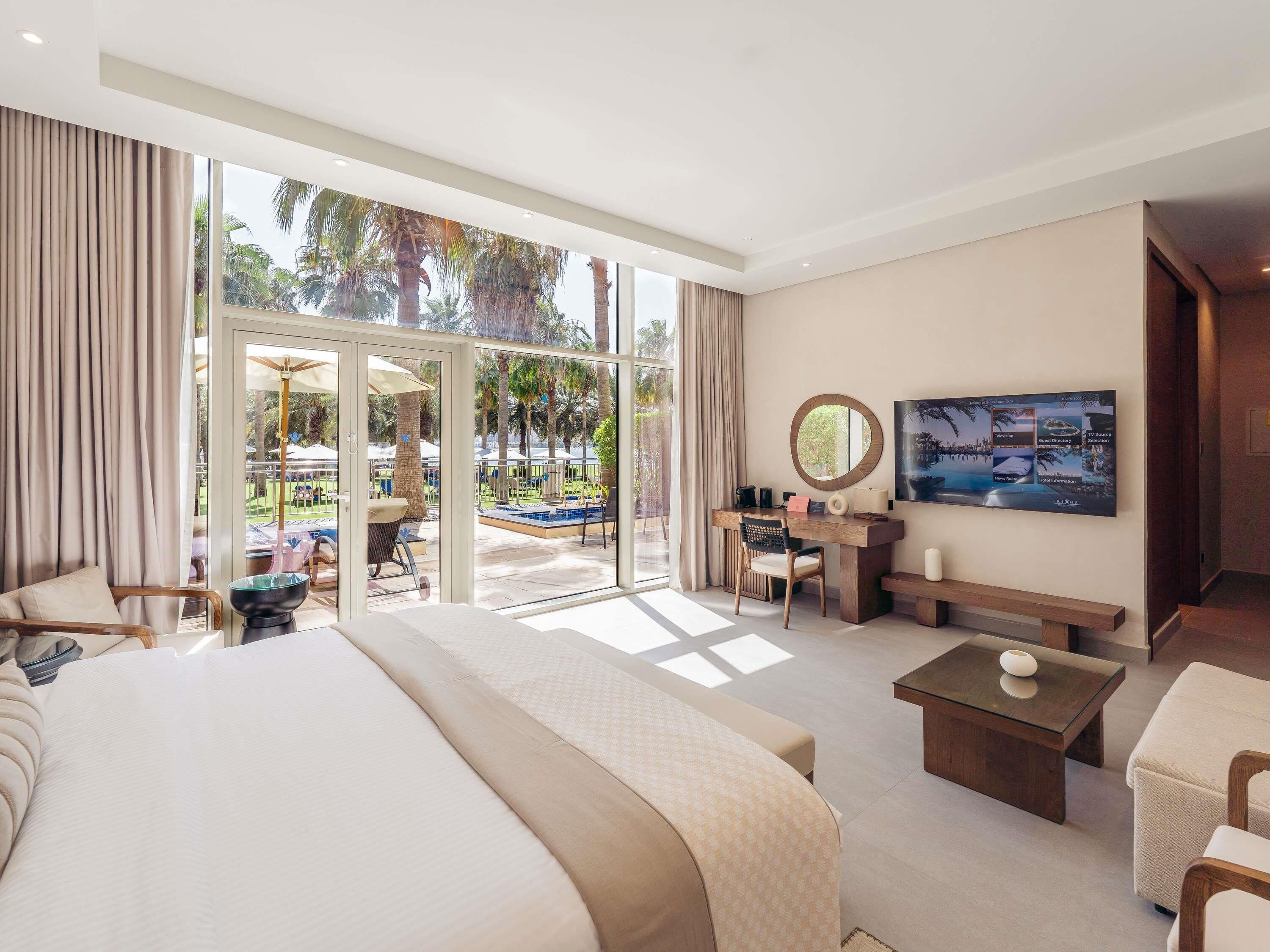 A spacious hotel room with a large bed, a desk, a TV, and a view of a pool and palm trees.