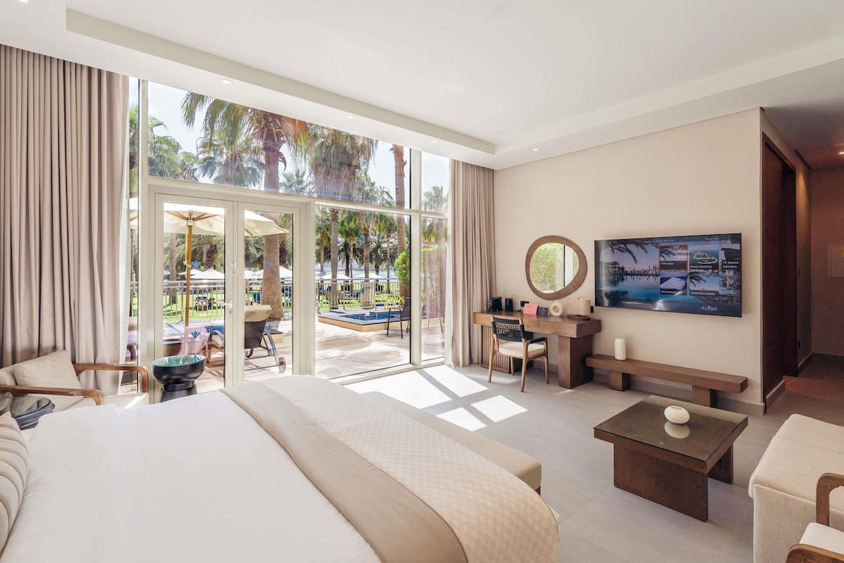 A spacious hotel room with a large bed, a desk, a TV, and a view of a pool and palm trees.