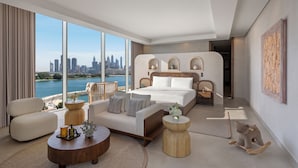A modern hotel room with a large bed, a sofa, a coffee table, and a view of a city skyline and water. - Rixos The Palm Hotel & Suites – Ultra All Inclusive (Dubai)
