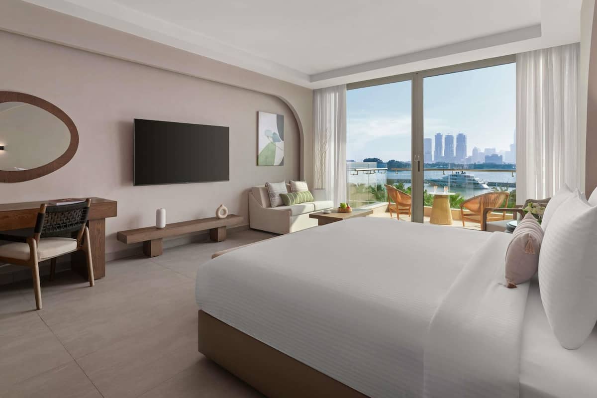 A modern hotel room with a large bed, a desk, a chair, a TV, and a balcony with a city view.