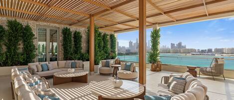 A spacious outdoor lounge area with multiple seating options, including sofas and chairs, arranged around a central coffee table. The area features a wooden pergola overhead and is adorned with green plants. A city skyline and waterfront are visible in the background.