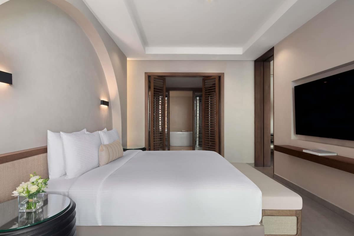 A modern hotel room with a large bed, a TV, and a glass side table with a vase of flowers.