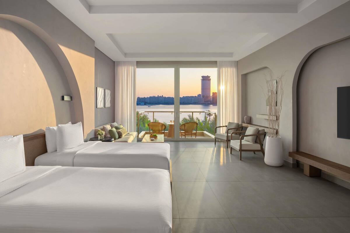 A hotel room with two beds, a balcony with a view of a city skyline and water, and a seating area with a table and chairs.