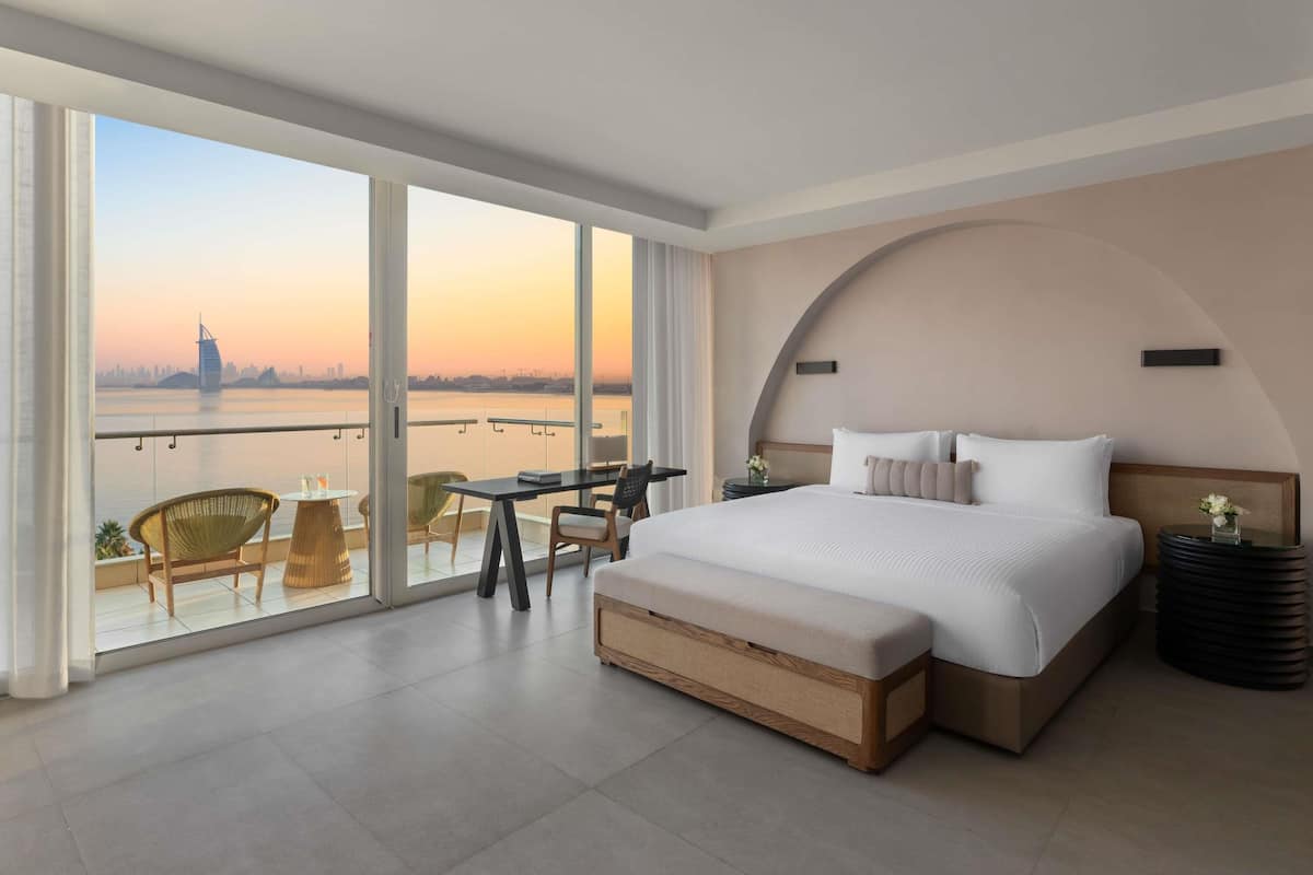 A modern hotel room with a large bed, a balcony with a view of a city skyline, and a dining area with chairs and a table.