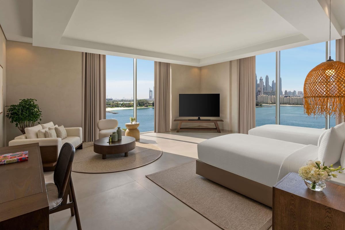 A modern hotel room with a large bed, a TV, a seating area with a coffee table, and a view of a city skyline and water.