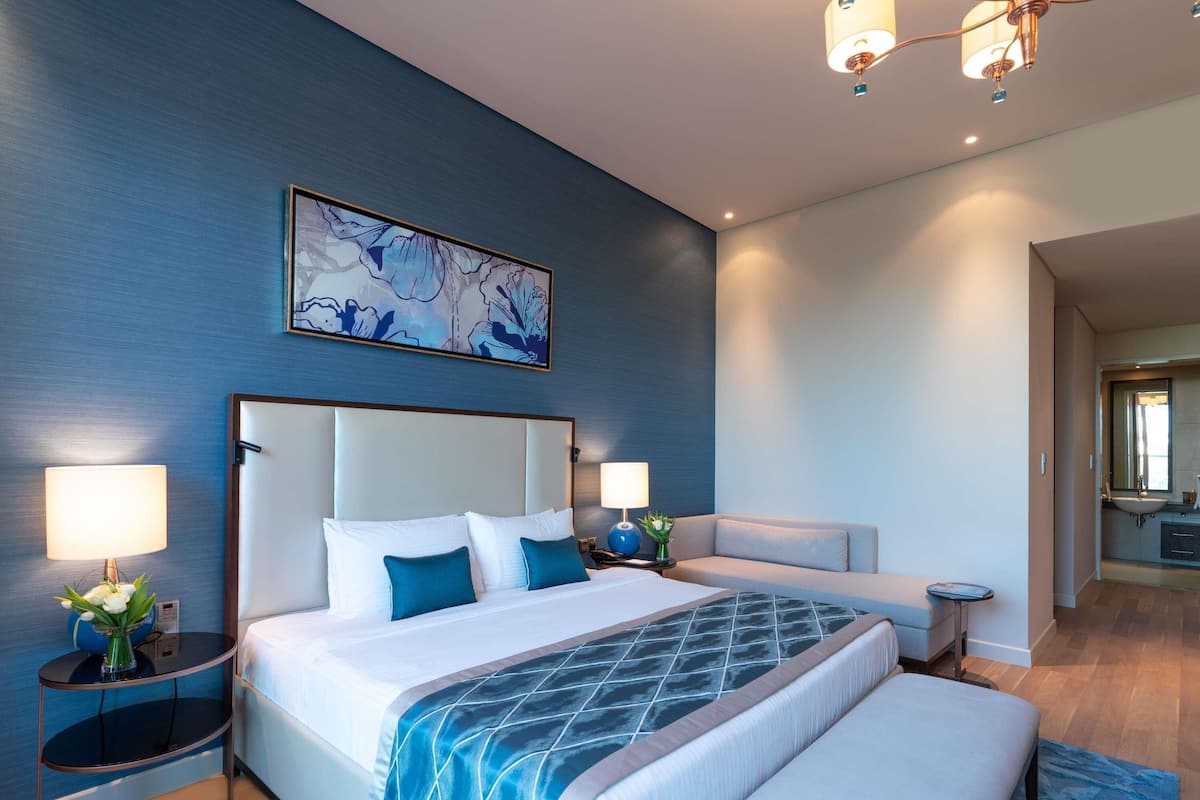 A modern hotel room with a large bed, two bedside tables, a sofa, and a bathroom visible through an open door.