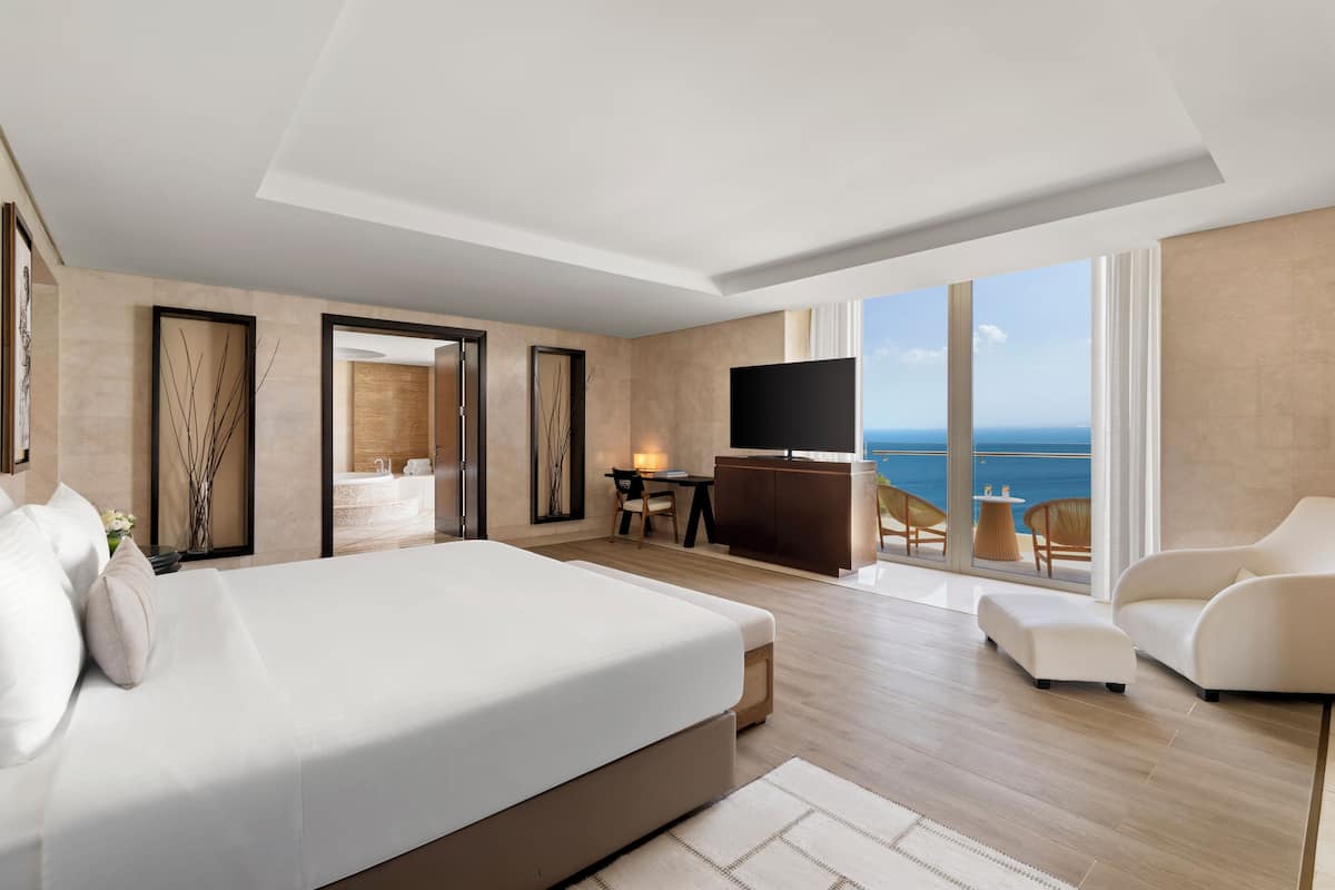 A modern hotel room with a large bed, a desk, a TV, and a balcony with a view of the sea.