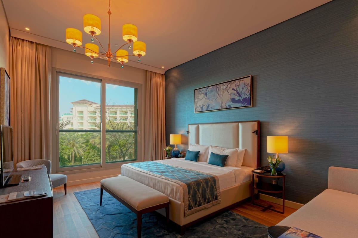 A hotel room with a large bed, a desk, a bench, a chair, a TV, a window with a view, and a chandelier.